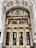 An ornate entrance of a historical building features a grand arched doorway with intricate carvings and decorative elements. The polished brass doors are reflective, showcasing a person taking a photo and the street behind them. Above the doors, the text 'United States Realty Building' is prominently displayed on an ornate arch.