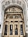 An ornate entrance of a historical building features a grand arched doorway with intricate carvings and decorative elements. The polished brass doors are reflective, showcasing a person taking a photo and the street behind them. Above the doors, the text 'United States Realty Building' is prominently displayed on an ornate arch.