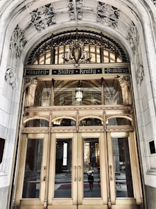 An ornate entrance of a historical building features a grand arched doorway with intricate carvings and decorative elements. The polished brass doors are reflective, showcasing a person taking a photo and the street behind them. Above the doors, the text 'United States Realty Building' is prominently displayed on an ornate arch.