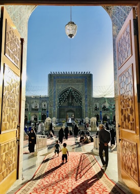 A grand architectural entrance opens onto a courtyard with a large ornate building in the background. The open wooden doors feature intricate geometric patterns. People, including women in traditional attire, walk and sit on a beautifully patterned red carpet leading towards the main structure. A crystal chandelier hangs above the open space.