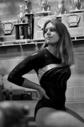 A person in a gymnastics leotard is sitting with a confident posture in front of a shelf filled with trophies and plaques. The room has a sporting ambiance with several awards displayed, indicating achievements.
