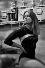A person in a gymnastics leotard is sitting with a confident posture in front of a shelf filled with trophies and plaques. The room has a sporting ambiance with several awards displayed, indicating achievements.
