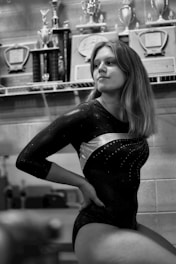 A person in a gymnastics leotard is sitting with a confident posture in front of a shelf filled with trophies and plaques. The room has a sporting ambiance with several awards displayed, indicating achievements.
