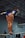 A gymnast in a blue leotard balances on a high bar in a gymnasium. The perspective is from below, highlighting the gymnast's focus and poise. The background shows a gym with a muted color palette and some gym equipment.