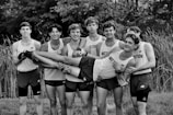 A group of seven young men wearing athletic uniforms, with 'Watterson' printed on their shirts. They are standing outdoors in front of tall grass. One of the young men is being held horizontally by the others, who are smiling and appearing to have a good time.