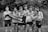 A group of seven young men wearing athletic uniforms, with 'Watterson' printed on their shirts. They are standing outdoors in front of tall grass. One of the young men is being held horizontally by the others, who are smiling and appearing to have a good time.