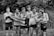 A group of seven young men wearing athletic uniforms, with 'Watterson' printed on their shirts. They are standing outdoors in front of tall grass. One of the young men is being held horizontally by the others, who are smiling and appearing to have a good time.