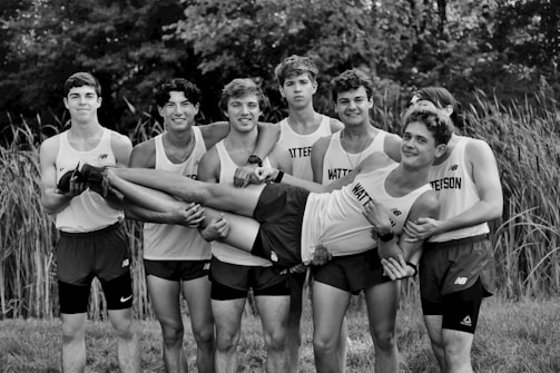 A group of seven young men wearing athletic uniforms, with 'Watterson' printed on their shirts. They are standing outdoors in front of tall grass. One of the young men is being held horizontally by the others, who are smiling and appearing to have a good time.