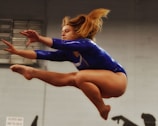 A gymnast performing a leap in mid-air with arms extended and knees bent, showcasing athleticism and grace. The gymnast is wearing a vibrant blue leotard, and the motion of her hair adds a dynamic element to the scene. The background is blurred, emphasizing the focus on the gymnast.