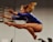 A gymnast performing a leap in mid-air with arms extended and knees bent, showcasing athleticism and grace. The gymnast is wearing a vibrant blue leotard, and the motion of her hair adds a dynamic element to the scene. The background is blurred, emphasizing the focus on the gymnast.
