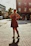 A smiling woman wearing a flowing red dress, standing in a sunlit urban street.