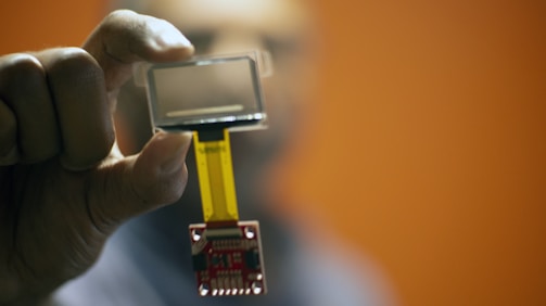 A close-up of a small electronic component with a transparent screen held between the thumb and forefinger of a person's hand. The background is blurred with a warm orange tone.