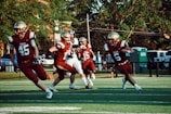 Football players wearing red and gold uniforms with the word 'Eagles' are actively running and playing on a field. The image captures a moment of action during daylight, surrounded by greenery and park or suburban setting with some parked vehicles and portable toilets in the background. The players are wearing helmets and are intensely focused on the game.