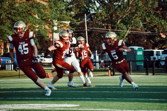 Football players wearing red and gold uniforms with the word 'Eagles' are actively running and playing on a field. The image captures a moment of action during daylight, surrounded by greenery and park or suburban setting with some parked vehicles and portable toilets in the background. The players are wearing helmets and are intensely focused on the game.