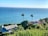 A coastal scene features two houses surrounded by lush greenery overlooking a vast, calm ocean with a clear blue sky above. Two tall palm trees stand prominently between the houses, adding a tropical feel to the landscape.