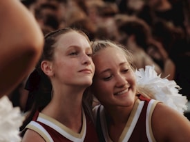 Two young women in cheerleader uniforms are standing closely together. One has a neutral expression while the other is smiling. They are surrounded by a crowd, suggesting a lively atmosphere.