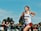 A young woman is running in a race, wearing athletic gear with a 'Watterson' logo and a bib number 2956. She appears focused and determined. In the background, a crowd of spectators is watching the event behind a fence. The sky is clear and blue, and there are trees behind the spectators.