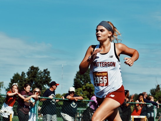 A young woman is running in a race, wearing athletic gear with a 'Watterson' logo and a bib number 2956. She appears focused and determined. In the background, a crowd of spectators is watching the event behind a fence. The sky is clear and blue, and there are trees behind the spectators.