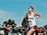 A young woman is running in a race, wearing athletic gear with a 'Watterson' logo and a bib number 2956. She appears focused and determined. In the background, a crowd of spectators is watching the event behind a fence. The sky is clear and blue, and there are trees behind the spectators.