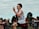 A young male athlete is captured mid-stride during a running event. He is wearing a white tank top, red shorts, and a race number pinned to his chest. Spectators and a fence line the background, with trees and a cloudy sky framing the scene.
