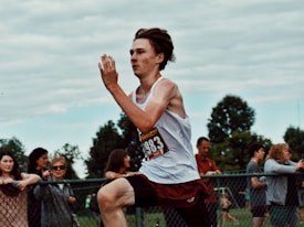 A young male athlete is captured mid-stride during a running event. He is wearing a white tank top, red shorts, and a race number pinned to his chest. Spectators and a fence line the background, with trees and a cloudy sky framing the scene.