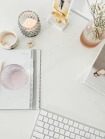 Flat lay of a minimalist workspace with a nude beige notebook, gold pen, and lavender-hued candles on a white desk.
