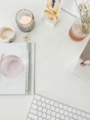 A minimalist workspace features a white desk with various items neatly arranged. A lit candle in a textured holder, a small vase with delicate white flowers, and a mug with a bracelet inside create a serene atmosphere. A spiral-bound planner is open to the month of February, and a small tile with an X mark rests on the desk. A keyboard is placed near the bottom of the image, indicating a workstation setup.