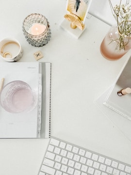 A minimalist workspace features a white desk with various items neatly arranged. A lit candle in a textured holder, a small vase with delicate white flowers, and a mug with a bracelet inside create a serene atmosphere. A spiral-bound planner is open to the month of February, and a small tile with an X mark rests on the desk. A keyboard is placed near the bottom of the image, indicating a workstation setup.