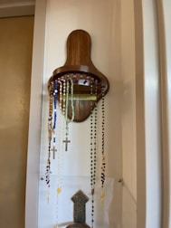 A wooden holder mounted on a wall displays several rosary beads in various colors, including blue, white, green, and purple. Each rosary is adorned with a crucifix. The holder has a simple, rustic design, and the wall is painted in a light color.