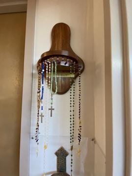 A wooden holder mounted on a wall displays several rosary beads in various colors, including blue, white, green, and purple. Each rosary is adorned with a crucifix. The holder has a simple, rustic design, and the wall is painted in a light color.