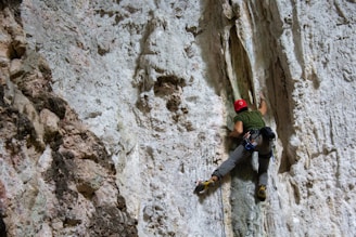 A person wearing climbing gear is scaling a steep, rocky cliff. The climber is using specialized equipment, including climbing shoes, harness, and ropes, to aid in their ascent. The rock surface is rough and textured, with various protrusions and crevices.