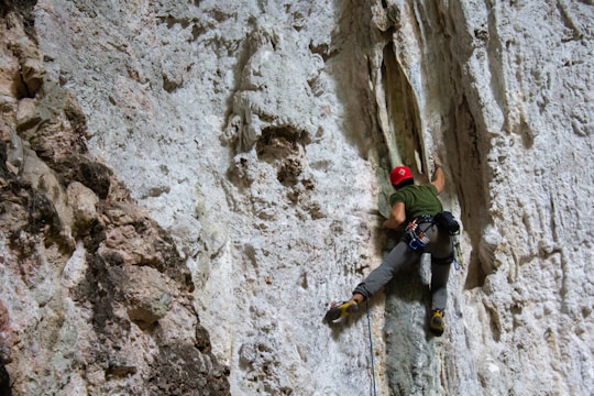 A person wearing climbing gear is scaling a steep, rocky cliff. The climber is using specialized equipment, including climbing shoes, harness, and ropes, to aid in their ascent. The rock surface is rough and textured, with various protrusions and crevices.