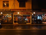 A dimly lit street scene at night featuring a bakery named Allan's Cake Hot Bread, a dry cleaning service, and a vintage store. The street is wet, suggesting recent rain. A person is walking on the sidewalk, partially visible and carrying an item. The shops have bright signs with various text, and light spills onto the street from shop windows.