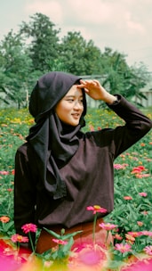 A woman wearing a dark hijab and clothing stands in the middle of a vibrant flower field. She is smiling and shading her eyes with her hand as she looks into the distance. The background features green foliage and trees, with a clear sky overhead.