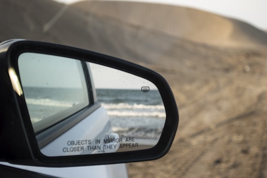 A car side mirror reflects the view of a sandy beach and ocean waves. The mirror is attached to a white vehicle, and contains the warning message 'Objects in mirror are closer than they appear.' The background out of the mirror shows sand dunes under a cloudy sky.