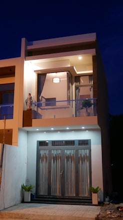 A modern two-story house with large glass doors on the ground floor and a balcony on the upper floor. The facade is well-lit and includes decorative plants and a statue to the left of the balcony area. The upper floor windows are framed by curtains, and there is a visible ceiling light within the interior. The house features a minimalist design with clean lines and contemporary architectural elements.