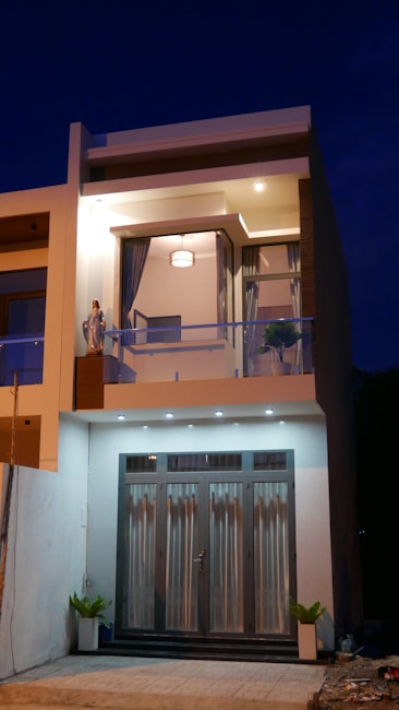 A modern two-story house with large glass doors on the ground floor and a balcony on the upper floor. The facade is well-lit and includes decorative plants and a statue to the left of the balcony area. The upper floor windows are framed by curtains, and there is a visible ceiling light within the interior. The house features a minimalist design with clean lines and contemporary architectural elements.