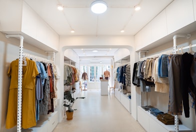 A well-organized clothing store with racks of assorted garments on both sides, featuring a mix of colors and styles. The space is brightly lit with white walls and a minimalist design, and a potted plant adds a touch of greenery.