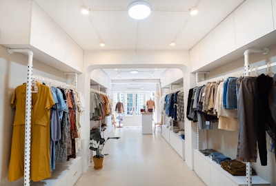 A well-organized clothing store with racks of assorted garments on both sides, featuring a mix of colors and styles. The space is brightly lit with white walls and a minimalist design, and a potted plant adds a touch of greenery.