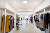 A well-organized clothing store with racks of assorted garments on both sides, featuring a mix of colors and styles. The space is brightly lit with white walls and a minimalist design, and a potted plant adds a touch of greenery.