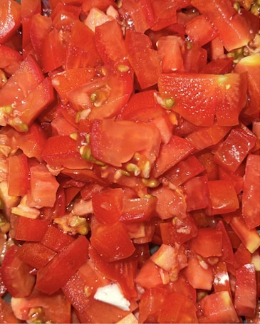Chopped pieces of fresh red tomatoes are closely packed together. The tomatoes are diced into various sizes, revealing their juicy interior with seeds visible. The vibrant red color dominates the image, with hints of green and white from seeds and tomato flesh.