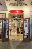 An open entrance to a store with wooden doors featuring glass panels and floral designs. The interior showcases a wall with a colorful display of items, possibly CDs or books, neatly arranged. The decor includes large, artistic floral patterns on the walls and ceiling, with a prominent red and black sign above the entrance reading 'dema3ors'. A staircase inside leads to an upper floor, adding depth to the cozy indoor ambiance.