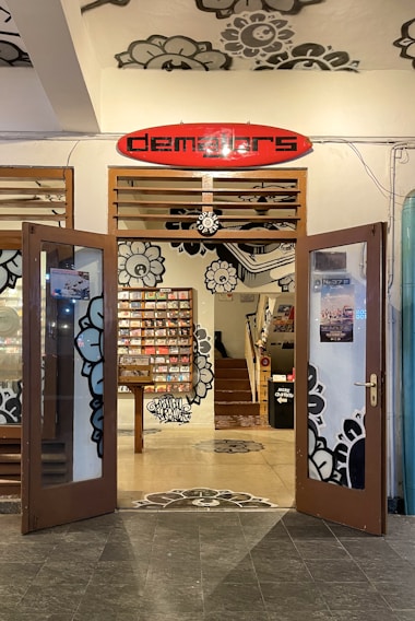 An open entrance to a store with wooden doors featuring glass panels and floral designs. The interior showcases a wall with a colorful display of items, possibly CDs or books, neatly arranged. The decor includes large, artistic floral patterns on the walls and ceiling, with a prominent red and black sign above the entrance reading 'dema3ors'. A staircase inside leads to an upper floor, adding depth to the cozy indoor ambiance.