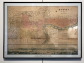 A framed vintage map of Shanghai is displayed on the wall. The map is detailed, showing various sections of the city and is colored with muted tones of reds, yellows, and browns. Labels and writing are in both English and possibly Chinese characters. A river flows through the map, labeled as 'POO RIVER.' Below the map is a panoramic illustration of Shanghai's skyline, featuring buildings and small boats on the water.