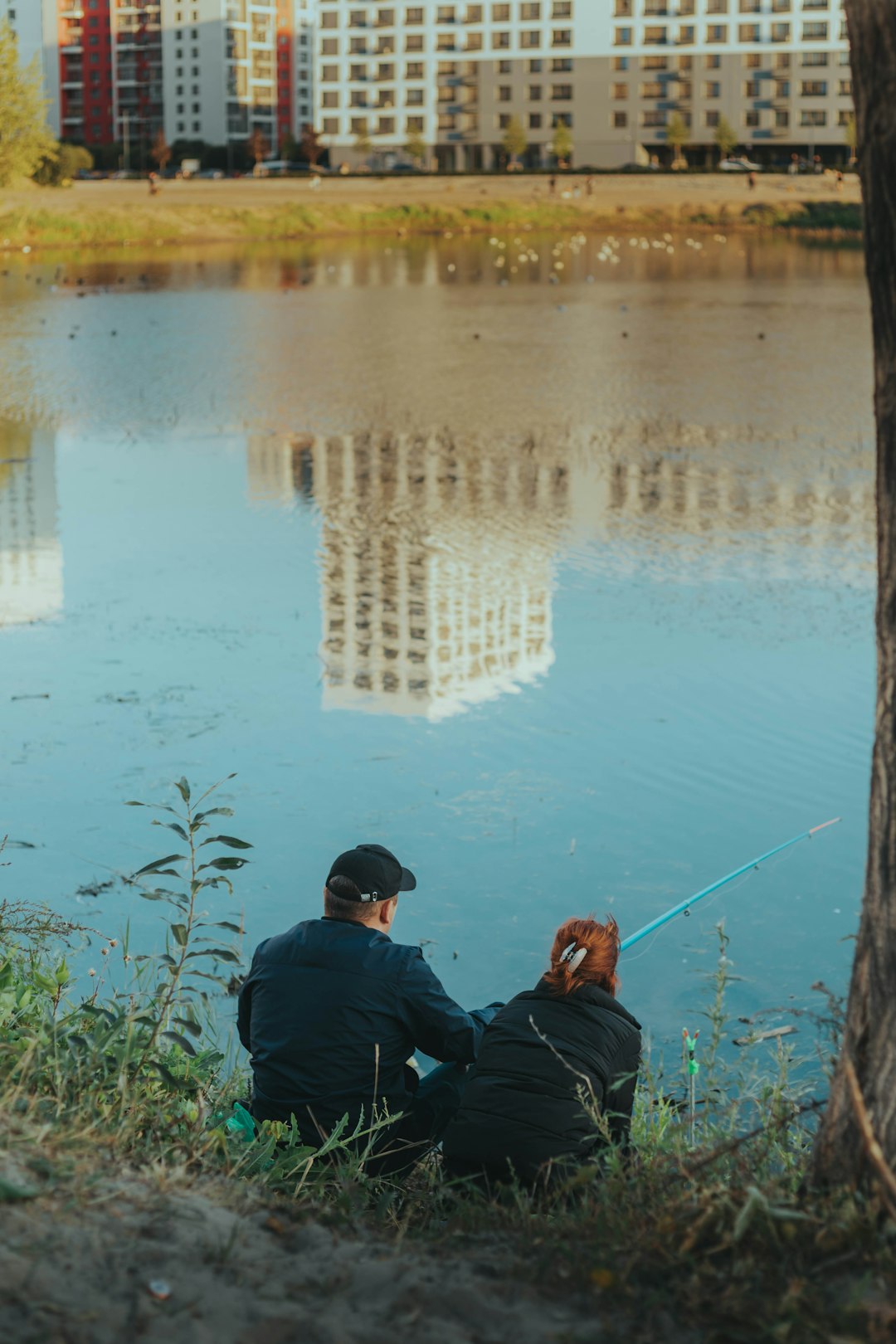 man in black jacket and blue denim jeans sitting on green grass near body of water