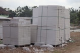 Large stacks of white concrete blocks are neatly arranged on wooden pallets. They are outside, beside a dirt path with some debris scattered around. Trees and a building with a corrugated metal roof are visible in the background.