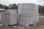 Large stacks of white concrete blocks are neatly arranged on wooden pallets. They are outside, beside a dirt path with some debris scattered around. Trees and a building with a corrugated metal roof are visible in the background.