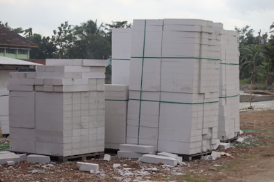 Large stacks of white concrete blocks are neatly arranged on wooden pallets. They are outside, beside a dirt path with some debris scattered around. Trees and a building with a corrugated metal roof are visible in the background.