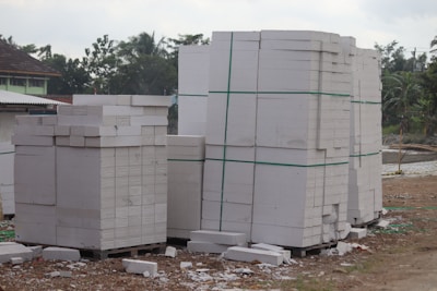 Large stacks of white concrete blocks are neatly arranged on wooden pallets. They are outside, beside a dirt path with some debris scattered around. Trees and a building with a corrugated metal roof are visible in the background.