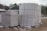 Large stacks of white concrete blocks are neatly arranged on wooden pallets. They are outside, beside a dirt path with some debris scattered around. Trees and a building with a corrugated metal roof are visible in the background.